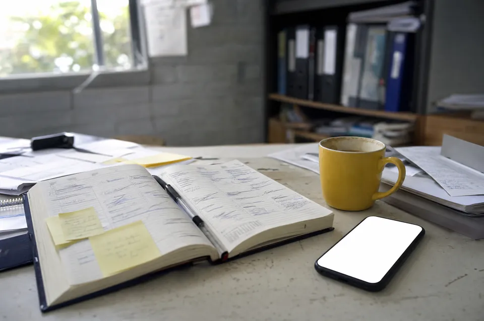 A messy paper diary full of crossed-out bookings, next to a coffee and a phone